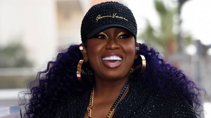 Missy Elliott smiles, wearing a studded cap, hoop earrings, and a gold necklace