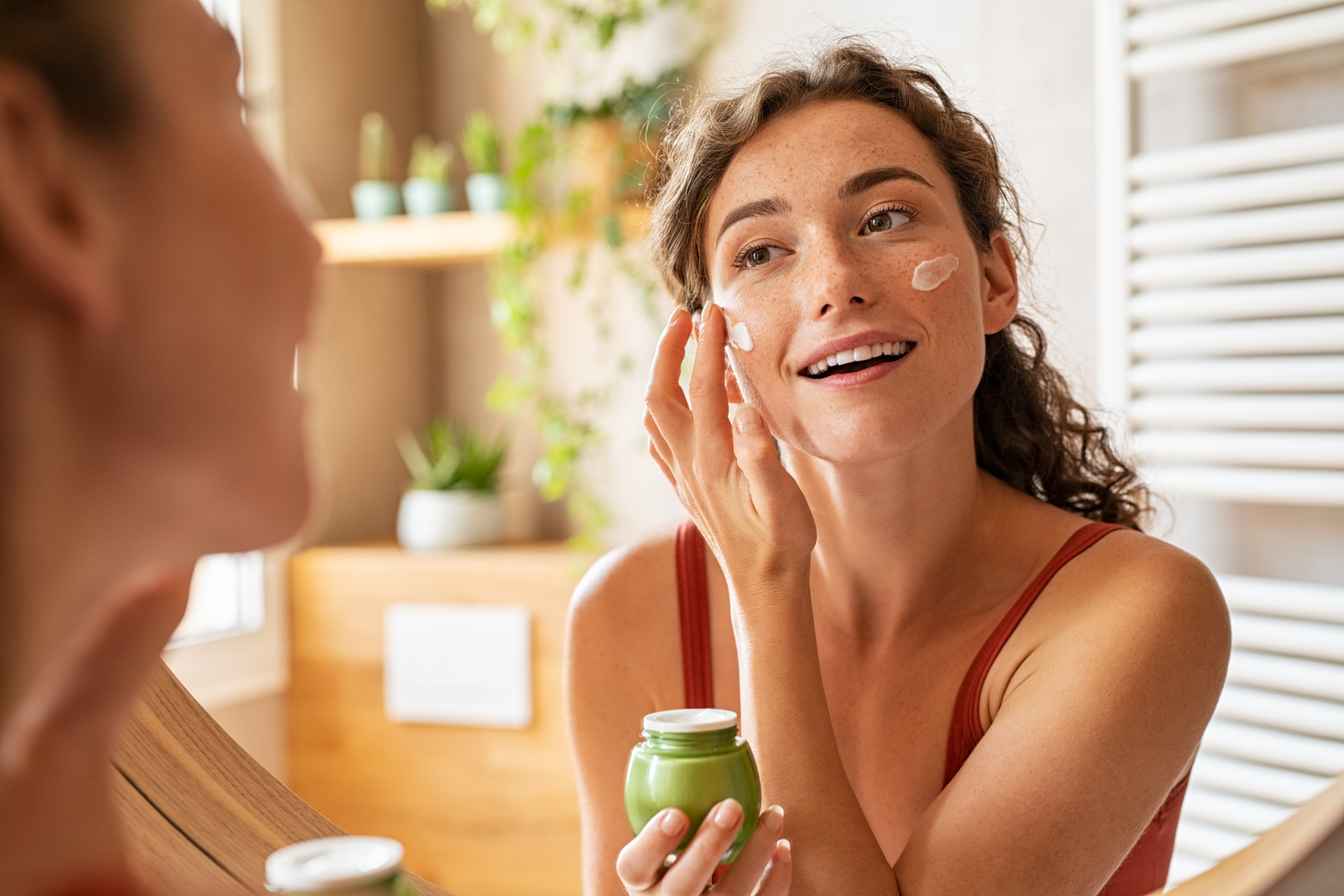 A woman in a bathroom applies cream to her face, holding a green jar, smiling at her reflection in the mirror