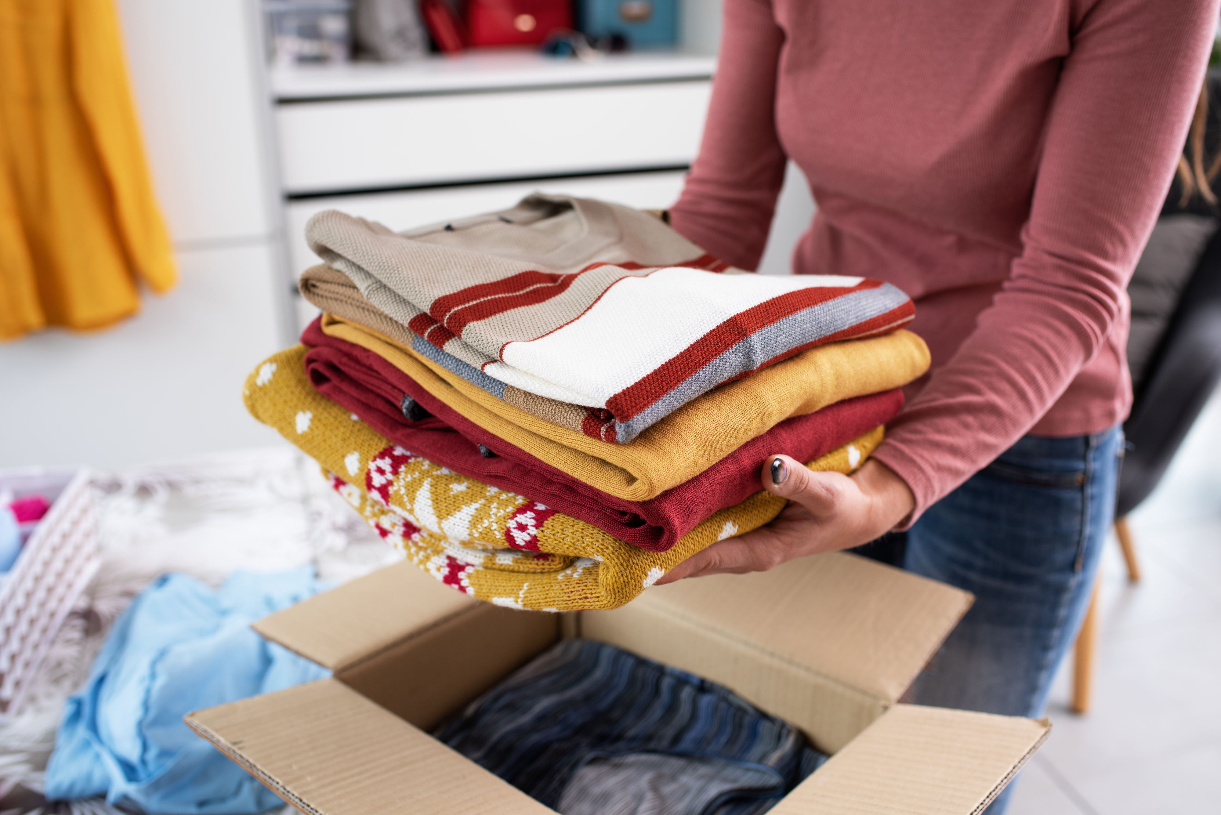 A person holds a stack of neatly folded clothes above an open cardboard box