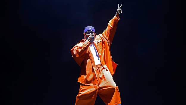 Tyler, The Creator performing on stage, wearing an oversized orange outfit and a purple cap, holding a microphone in one hand and raising the other in a peace sign