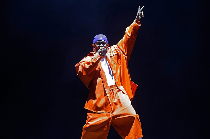 Tyler, The Creator performing on stage, wearing an oversized orange outfit and a purple cap, holding a microphone in one hand and raising the other in a peace sign