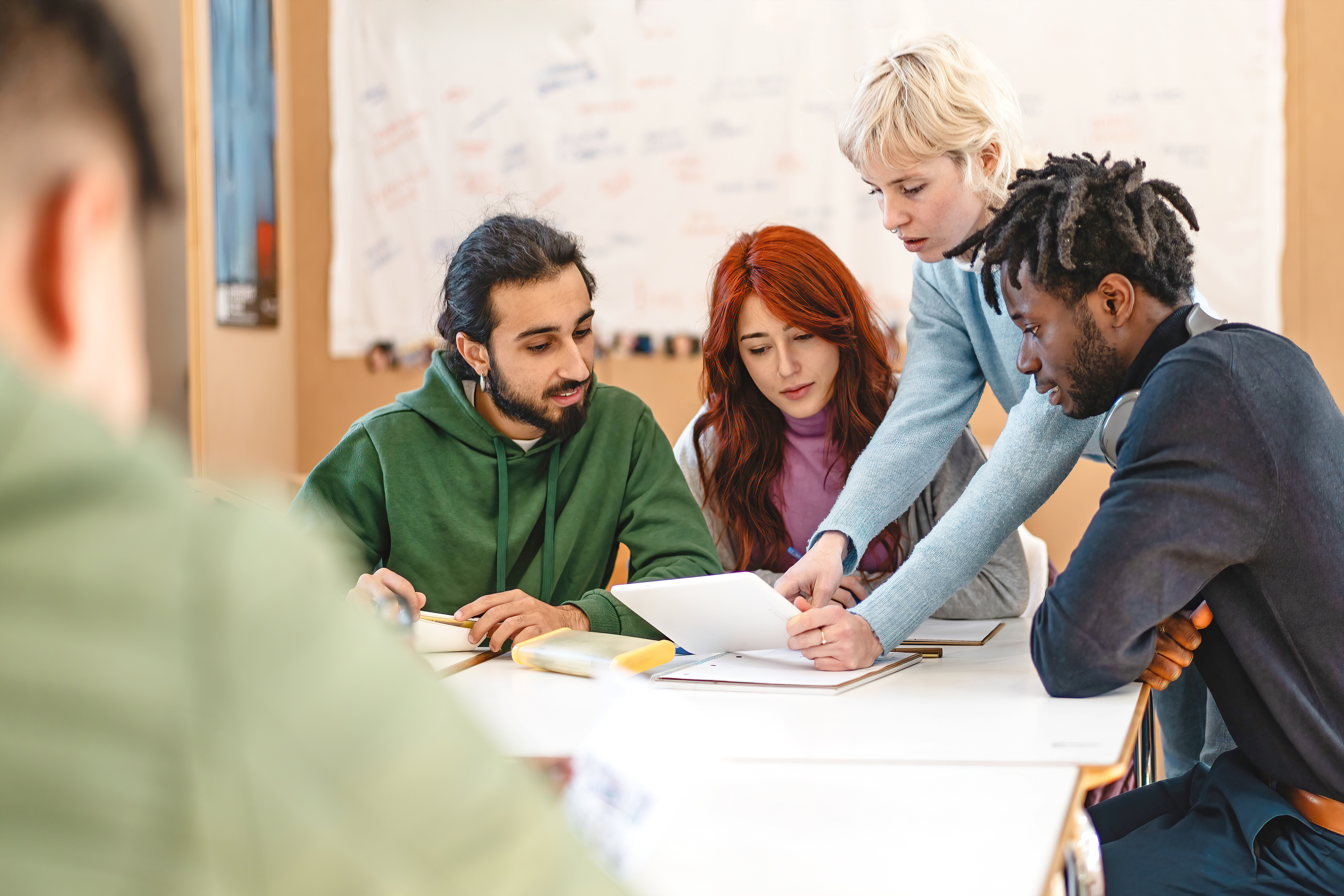 A group of four people, one directing attention to a paper, collaborates around a table with notes on a wall in the background