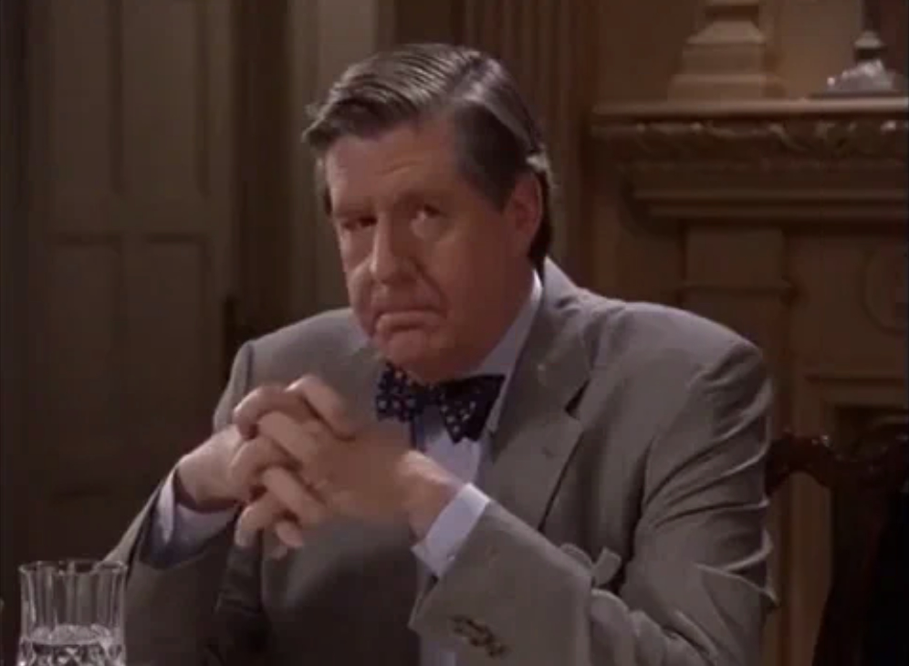 Older man wearing a suit with a bow tie, seated at a table with a serious expression