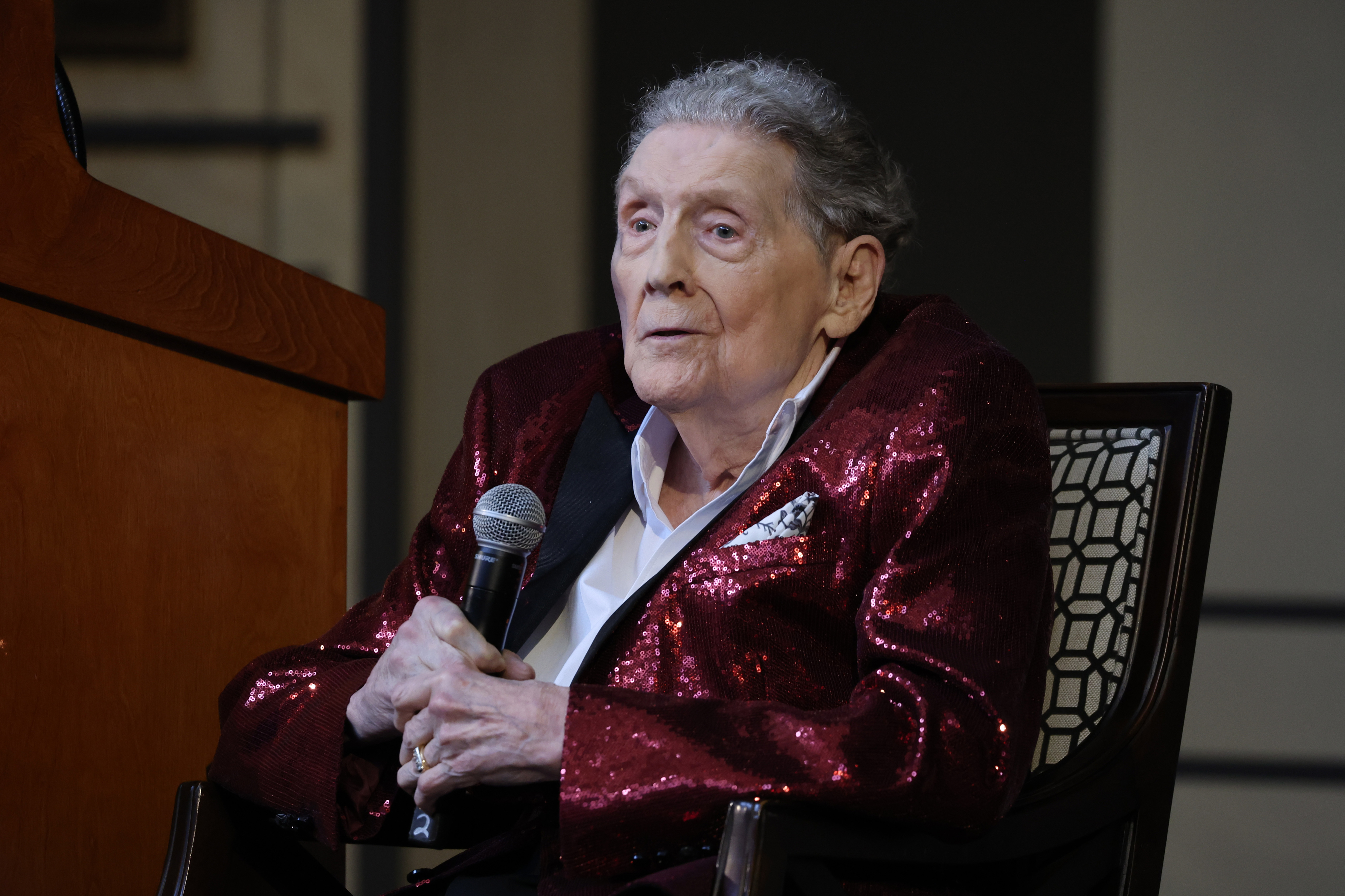 Elderly man in a sequin jacket holding a microphone while seated