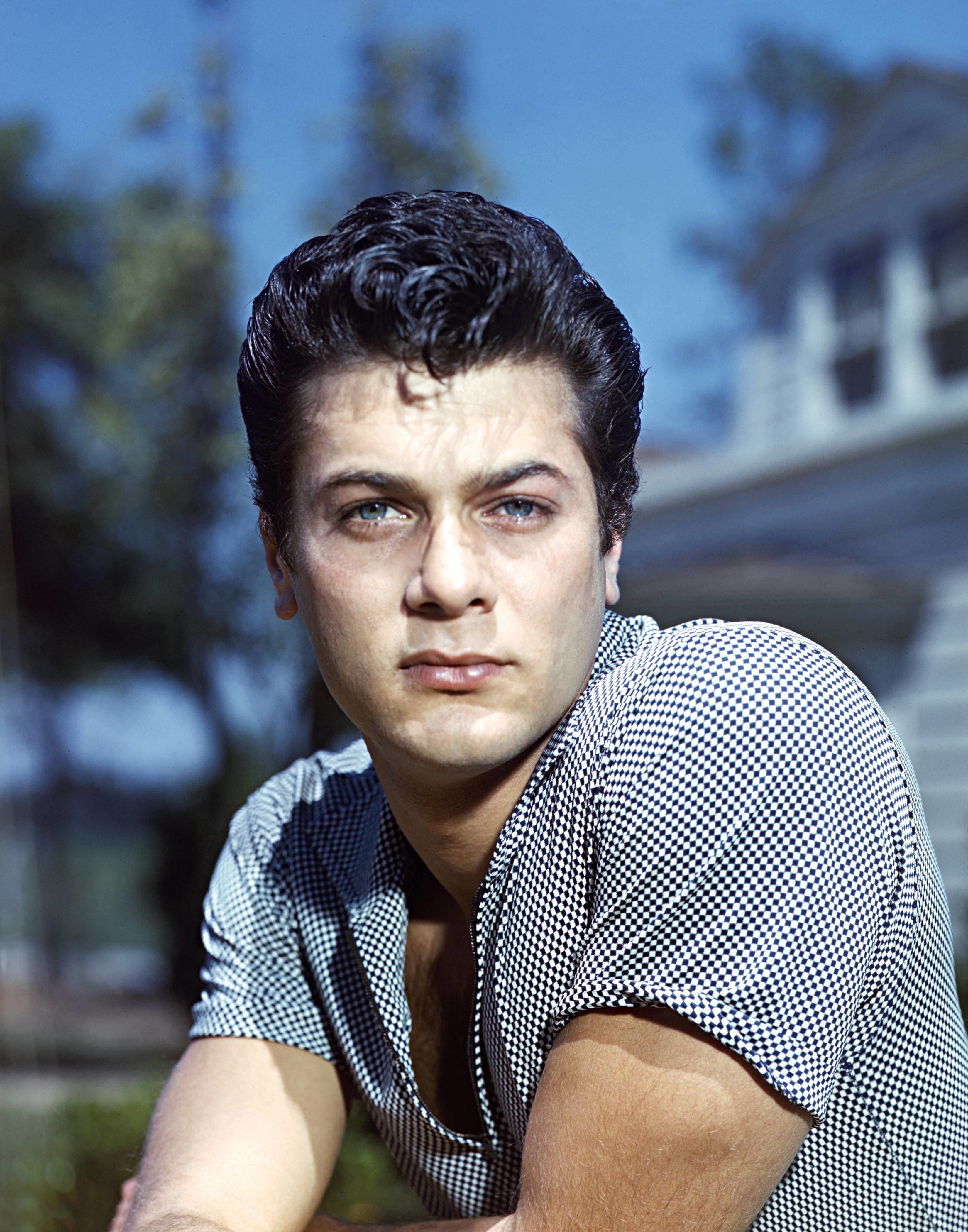 Tony Curtis with styled dark hair, wearing a checkered shirt, in a contemplative pose outdoors with trees and a house in the background