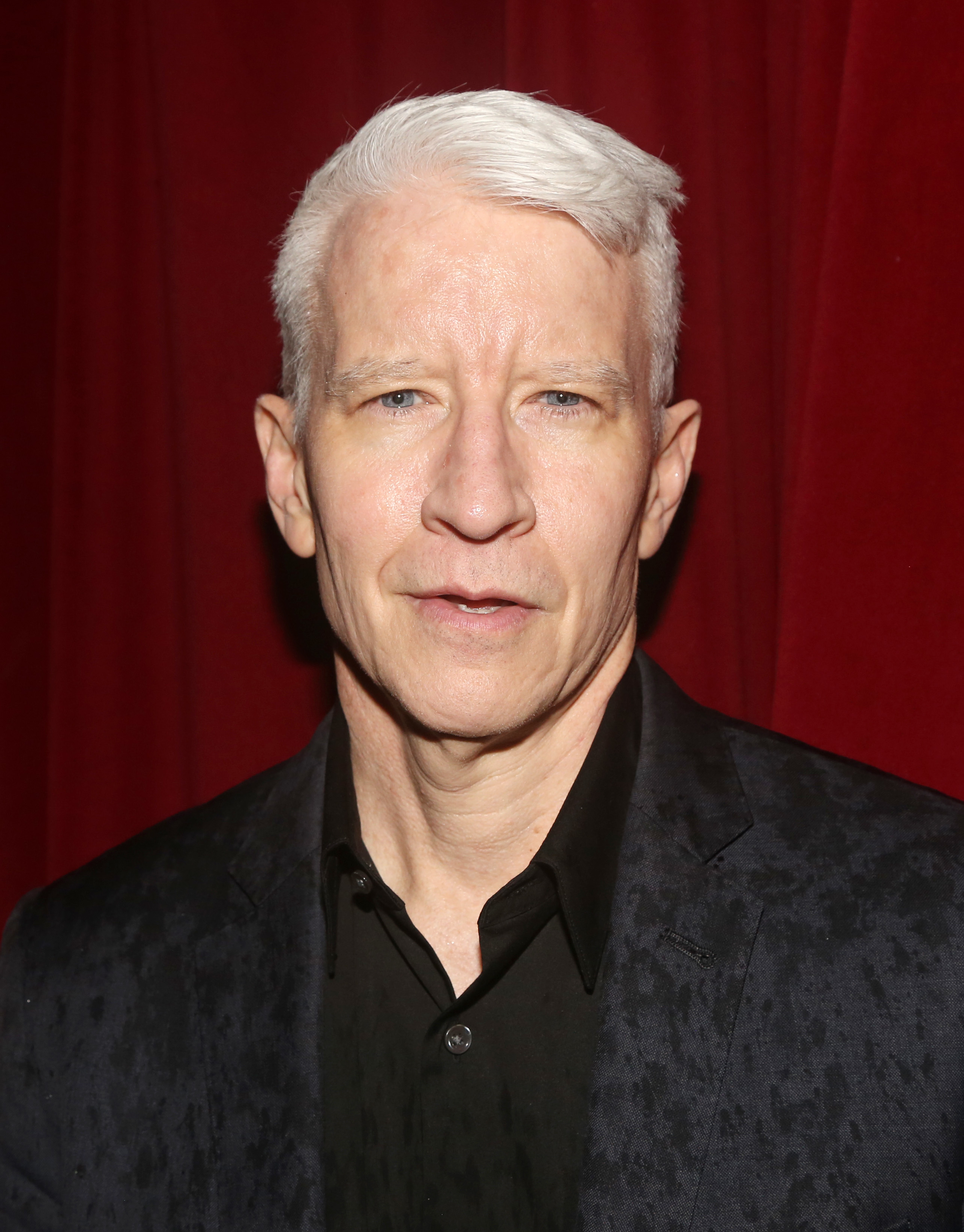 Anderson Cooper in a formal black suit and shirt, posing in front of a red curtain