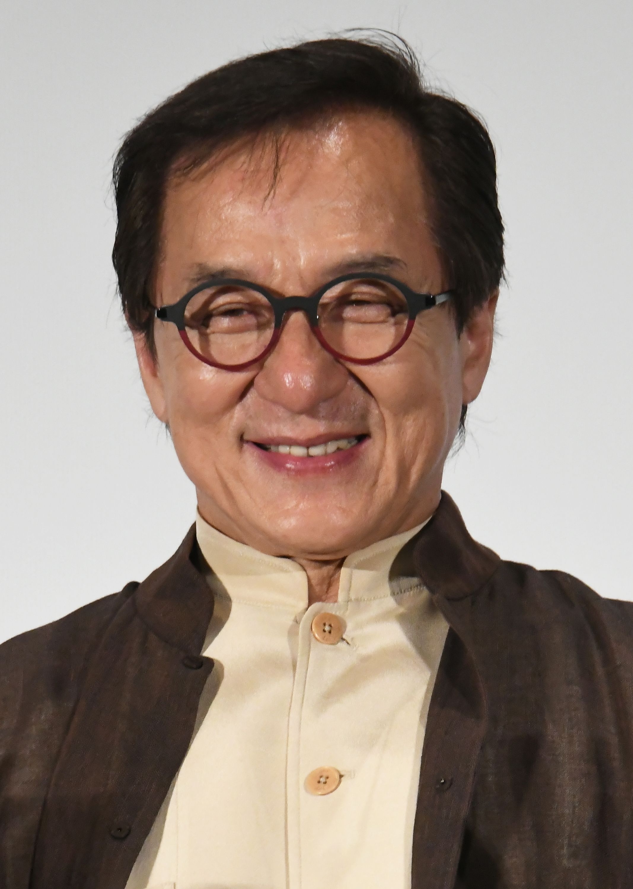 Jackie Chan smiling, wearing glasses and a traditional button-up outfit