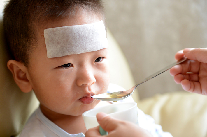 幼い男の子が額に冷却シートを貼り、スプーンで薬を飲んでいます。