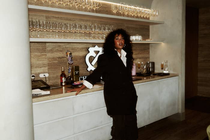 SZA stands in a chic, modern room with shelves of glasses and a champagne sign in the background. She wears a stylish, oversized coat
