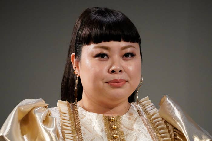 金髪のピアスを着けた女性が、ゴールドの豪華なドレスを着て、カメラを見ている。