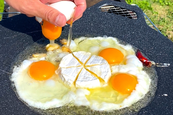 屋外の鉄板で、4つの目玉焼きとカマンベールチーズを溶かしながら焼いている様子。手が卵を割っているところ。