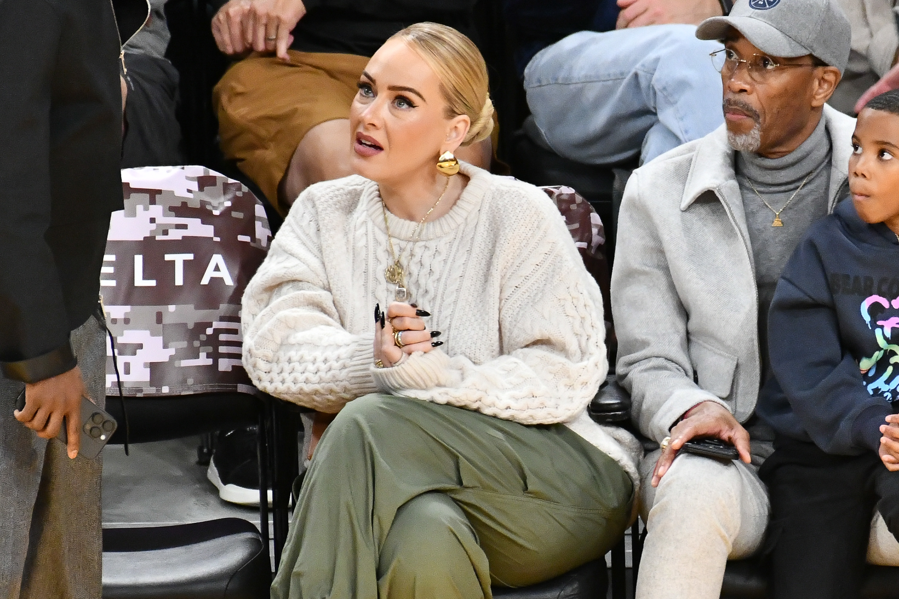 Adele sits courtside at a basketball game, wearing a cozy sweater and green pants. She is speaking to someone off-camera