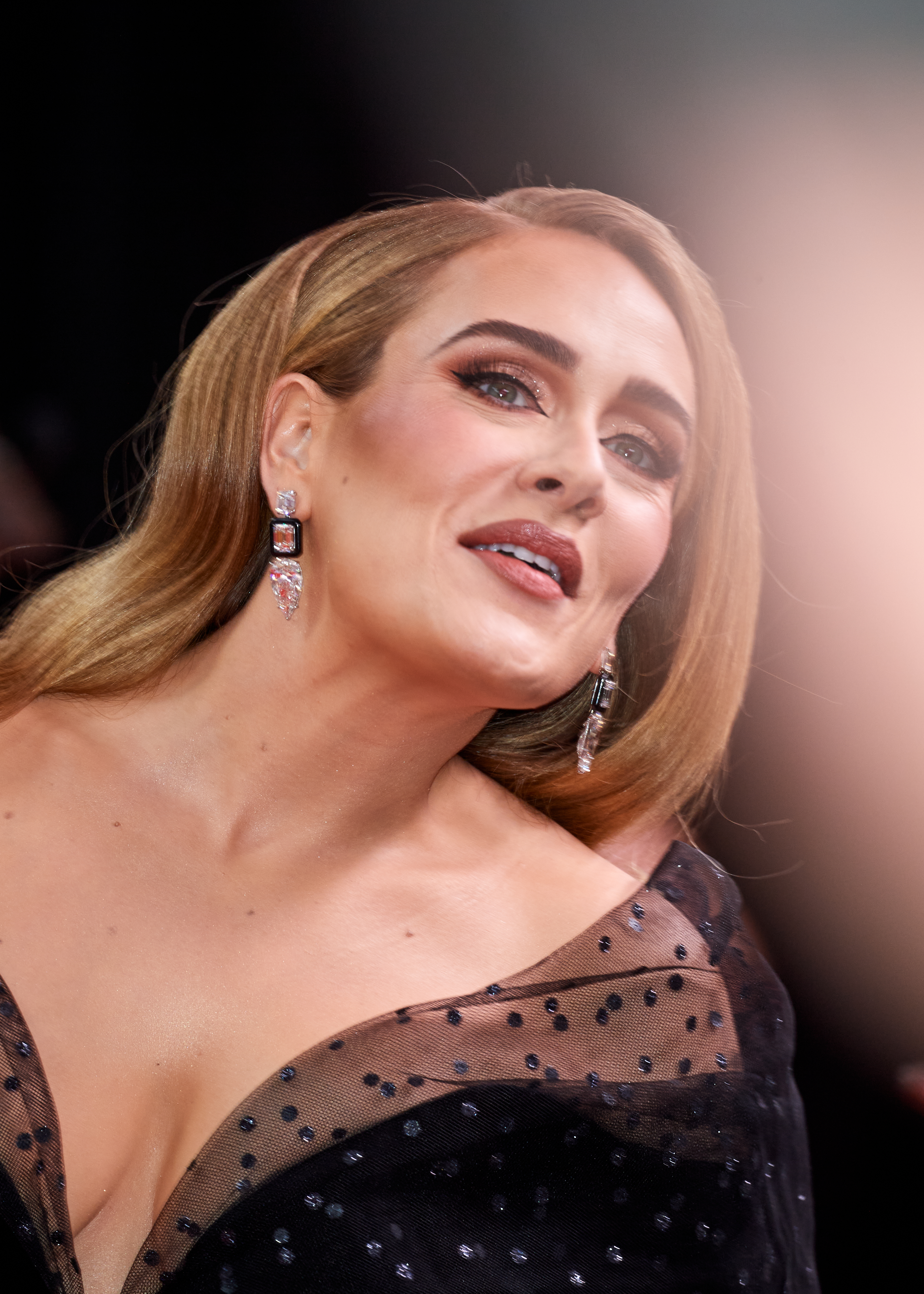 Adele on the red carpet in an elegant dress with sheer polka-dot fabric and statement earrings. She is posing with a slight smile