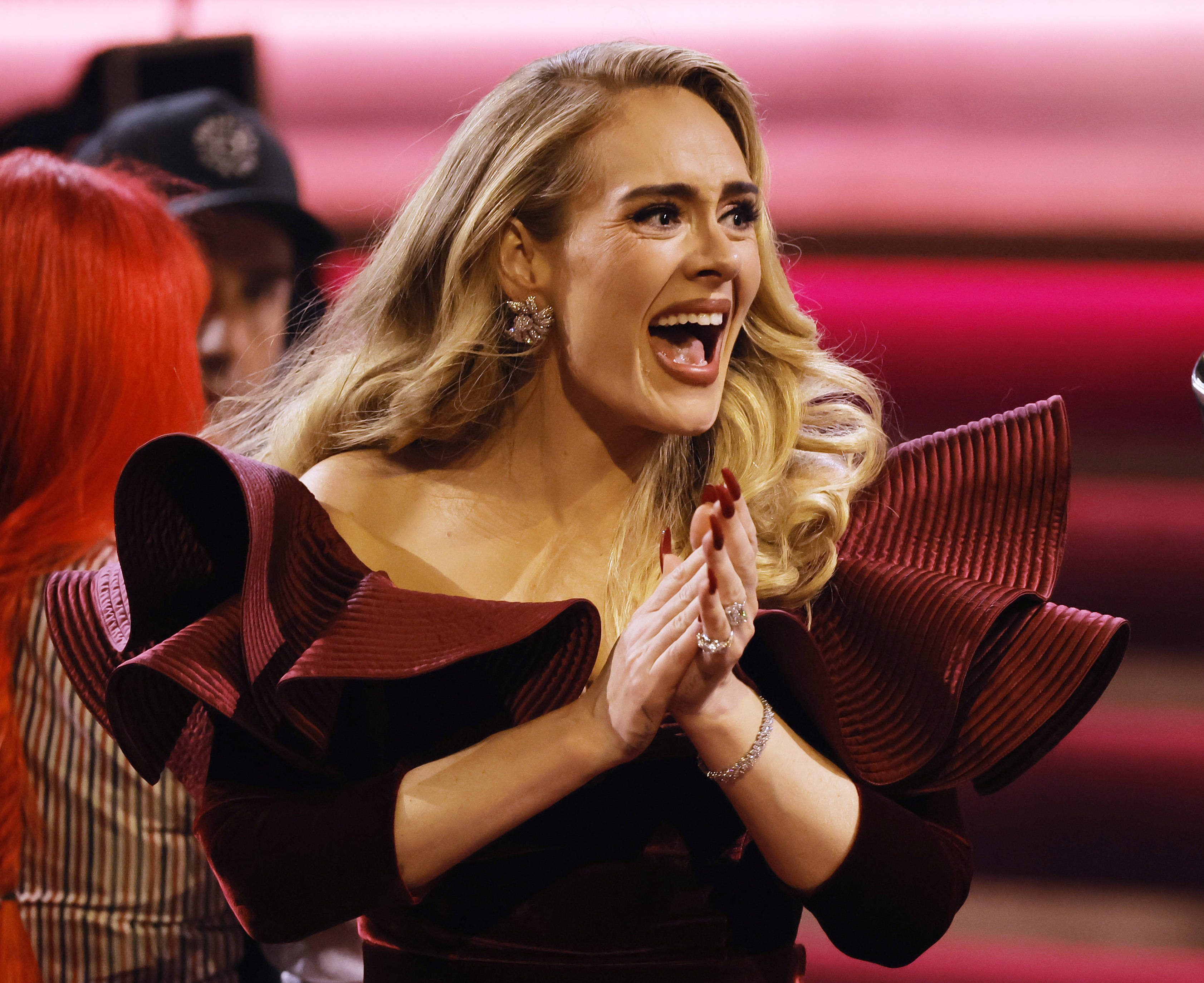 Adele, wearing an elegant, off-the-shoulder dress with large, ruffled sleeves, claps her hands and smiles excitedly