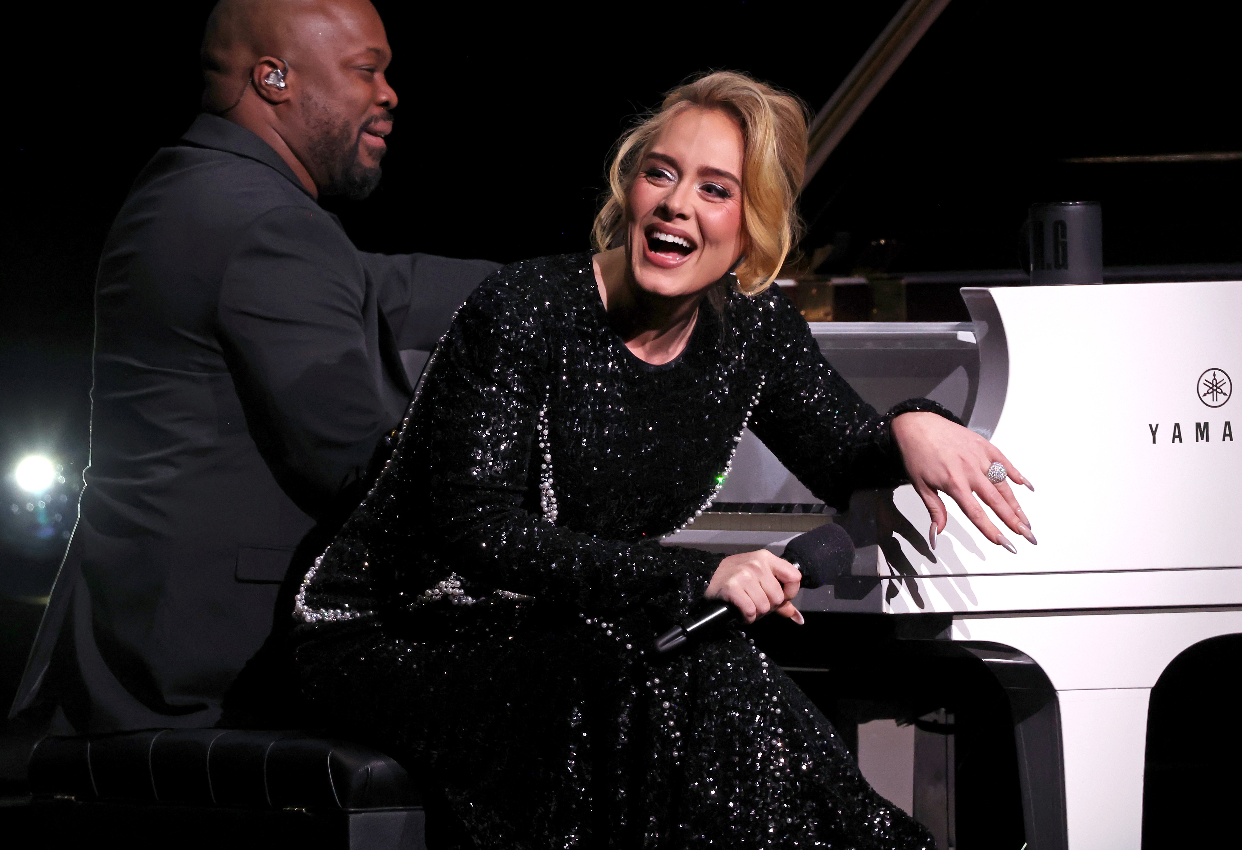 Adele, in a sparkly black gown, leans on a white Yamaha piano while smiling. A pianist, dressed in a dark suit, sits beside her during a performance