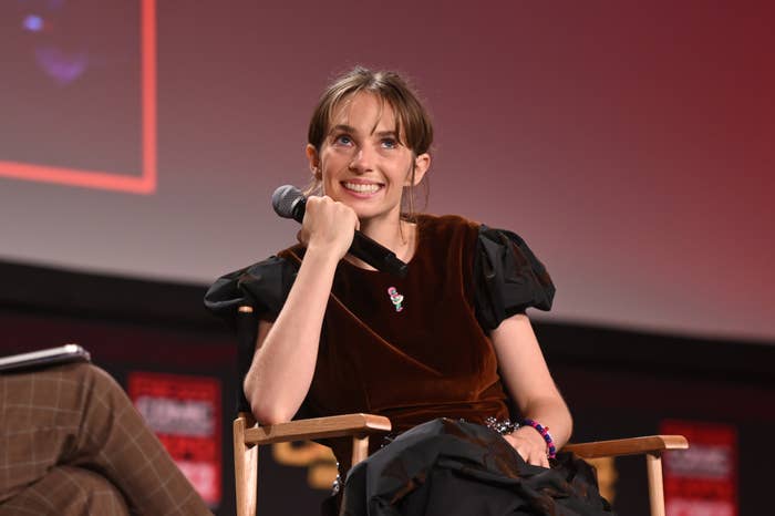 Maya Hawke sits on a stage during an event, holding a microphone and smiling. She wears a dark, puff-sleeve dress