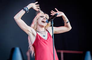 Woman singing energetically on stage, wearing a sleeveless dress, multiple necklaces, and wristbands