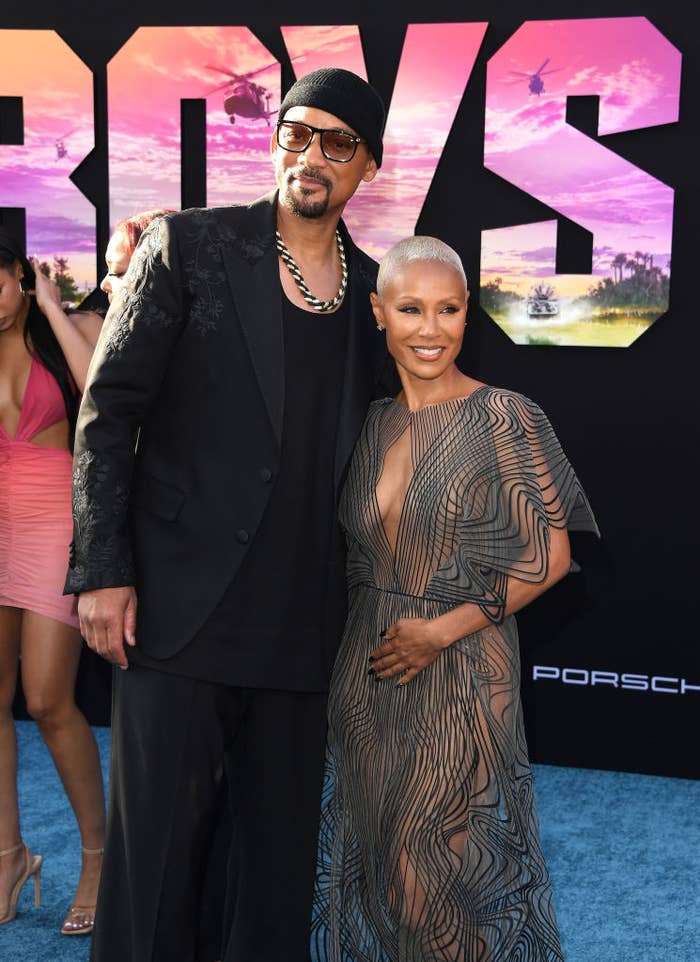 Will Smith in a black suit and Jada Pinkett Smith in a sheer, patterned dress, smiling together at the red carpet event for their movie