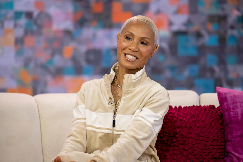 Jada Pinkett Smith sits on a couch, smiling, wearing a casual zip-up jacket. Bright background and pillows