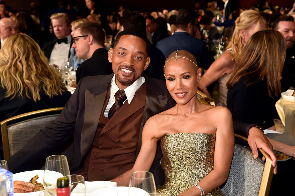 Will Smith and Jada Pinkett Smith smiling at a formal event, seated among other guests. Will is in a brown three-piece suit, Jada in a strapless sparkly dress