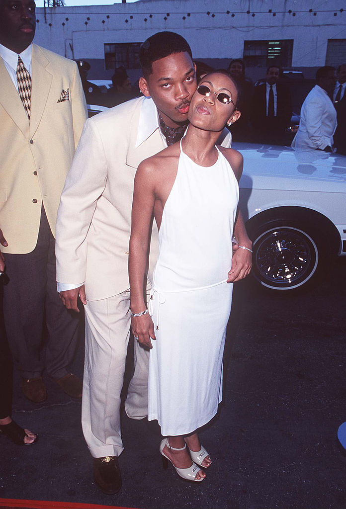 Will Smith kisses Jada Pinkett Smith on the head. Will wears a light suit, Jada wears a sleeveless dress. They are on a red carpet surrounded by people