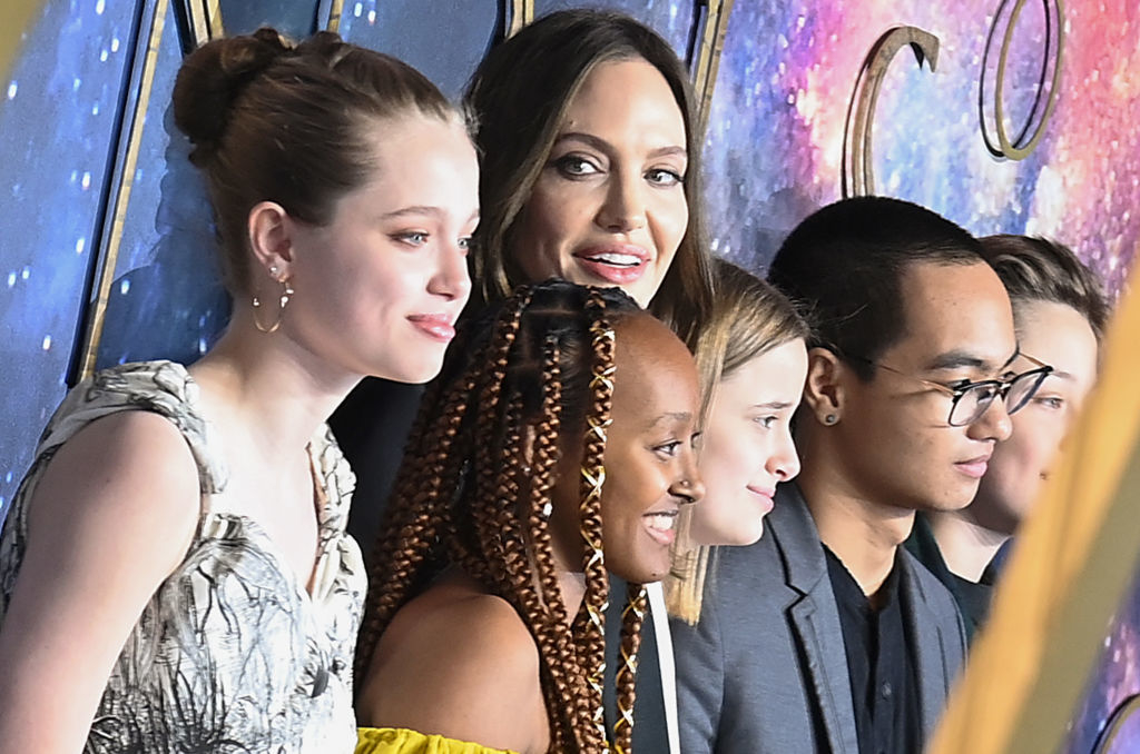 Angelina Jolie with Shiloh, Zahara, and Pax Jolie-Pitt posing together at an event. Shiloh in a patterned dress, Zahara in yellow, Pax in a black top and blazer