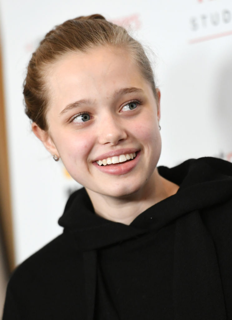 Shiloh Jolie-Pitt smiles with a casual hairstyle, wearing a simple black hoodie at a celebrity event backdrop