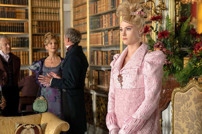 A polite young lady in a textured gown stands in a library with ornate decor. In the background, more guests converse, all in Regency-era attire