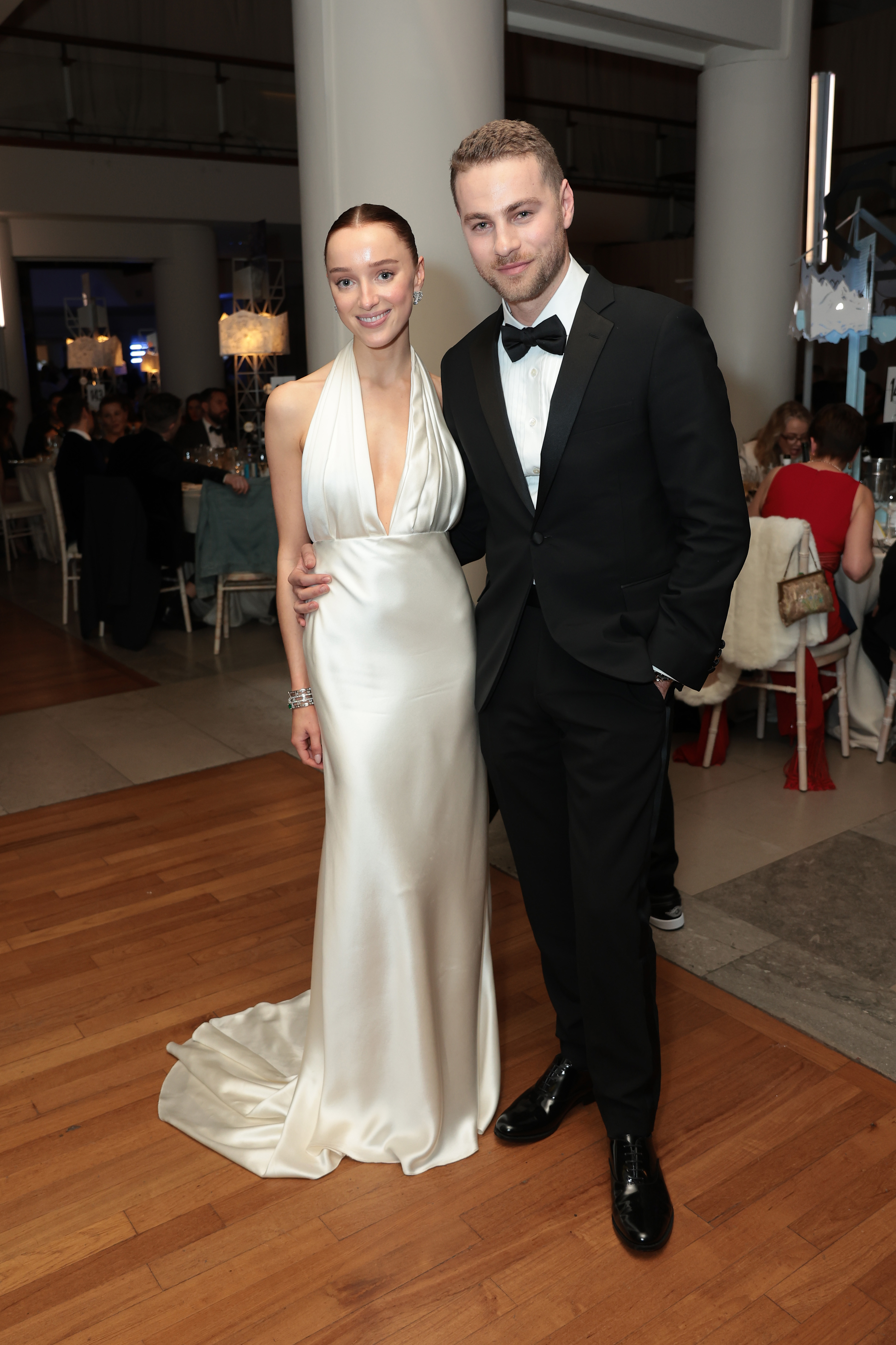 Phoebe Dynevor in a sleek, white halter-neck gown and Cameron Fuller in a black tuxedo, posing together at a formal event