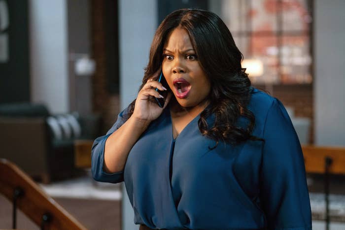 Amber Riley, in a casual outfit, holding a phone with a shocked expression