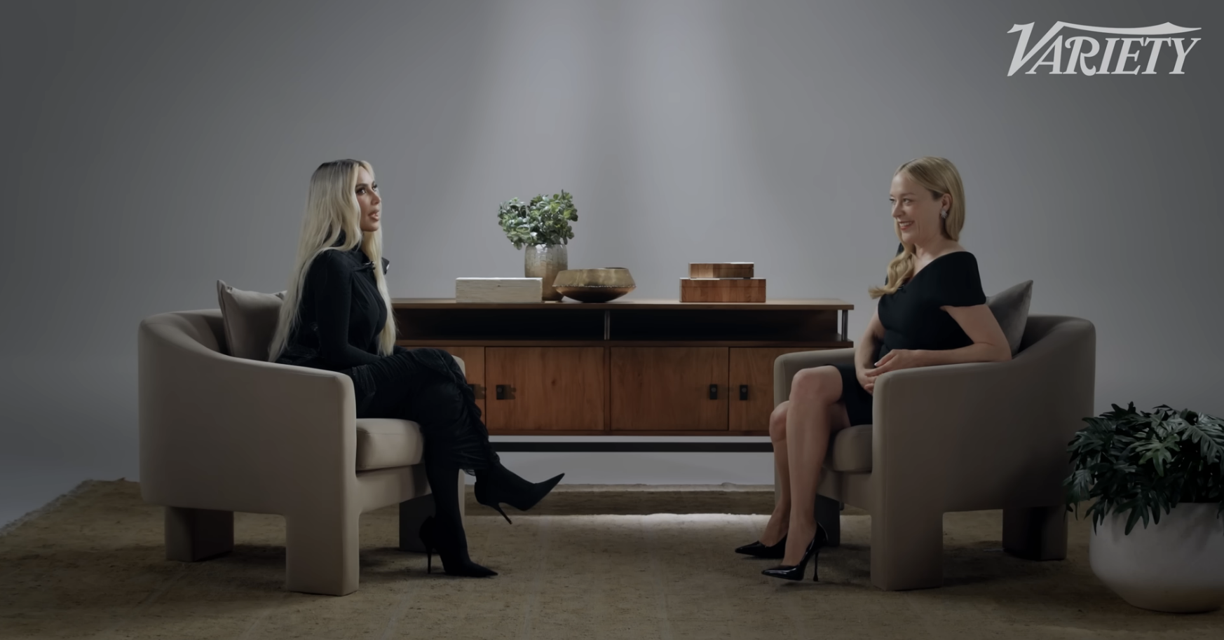 Kim Kardashian in dark outfit and boots, sits on a chair across from a smiling Chloë Sevigny in a dark dress for a Variety interview