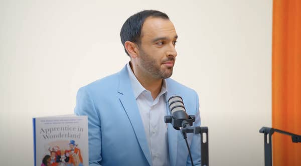 Ramin Setoodeh, in a light blazer, is sitting in front of a microphone. A book titled &quot;Apprentice in Wonderland&quot; is visible next to him
