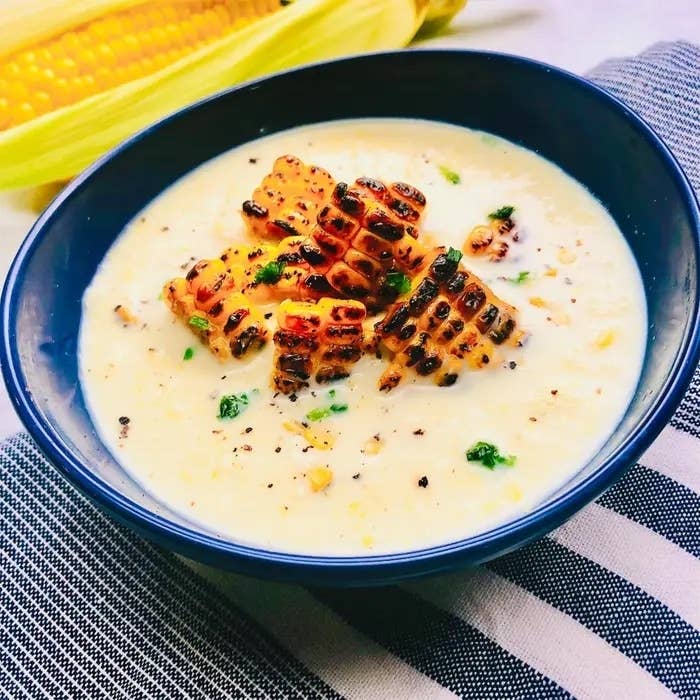 焼きトウモロコシのトッピングがのったコーンスープのボウル。背景にトウモロコシが見える。