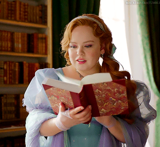Nicola Coughlan, in a historical gown with puffed sleeves, holds and reads an ornate book in a library setting with bookshelves in the background