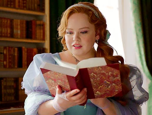 Nicola Coughlan, in a historical gown with puffed sleeves, holds and reads an ornate book in a library setting with bookshelves in the background