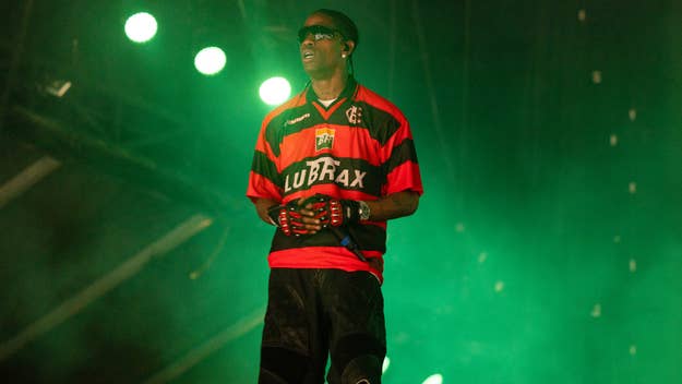 Travis Scott performs on stage, wearing a red and black soccer jersey, protective gloves, and sunglasses. Stage lights shine behind him