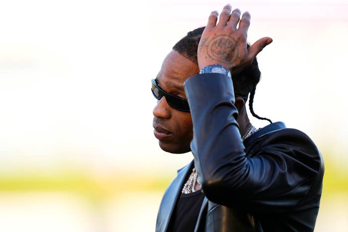 Travis Scott, wearing a dark leather jacket, dark shirt, and sunglasses, touching his hair while looking to the side