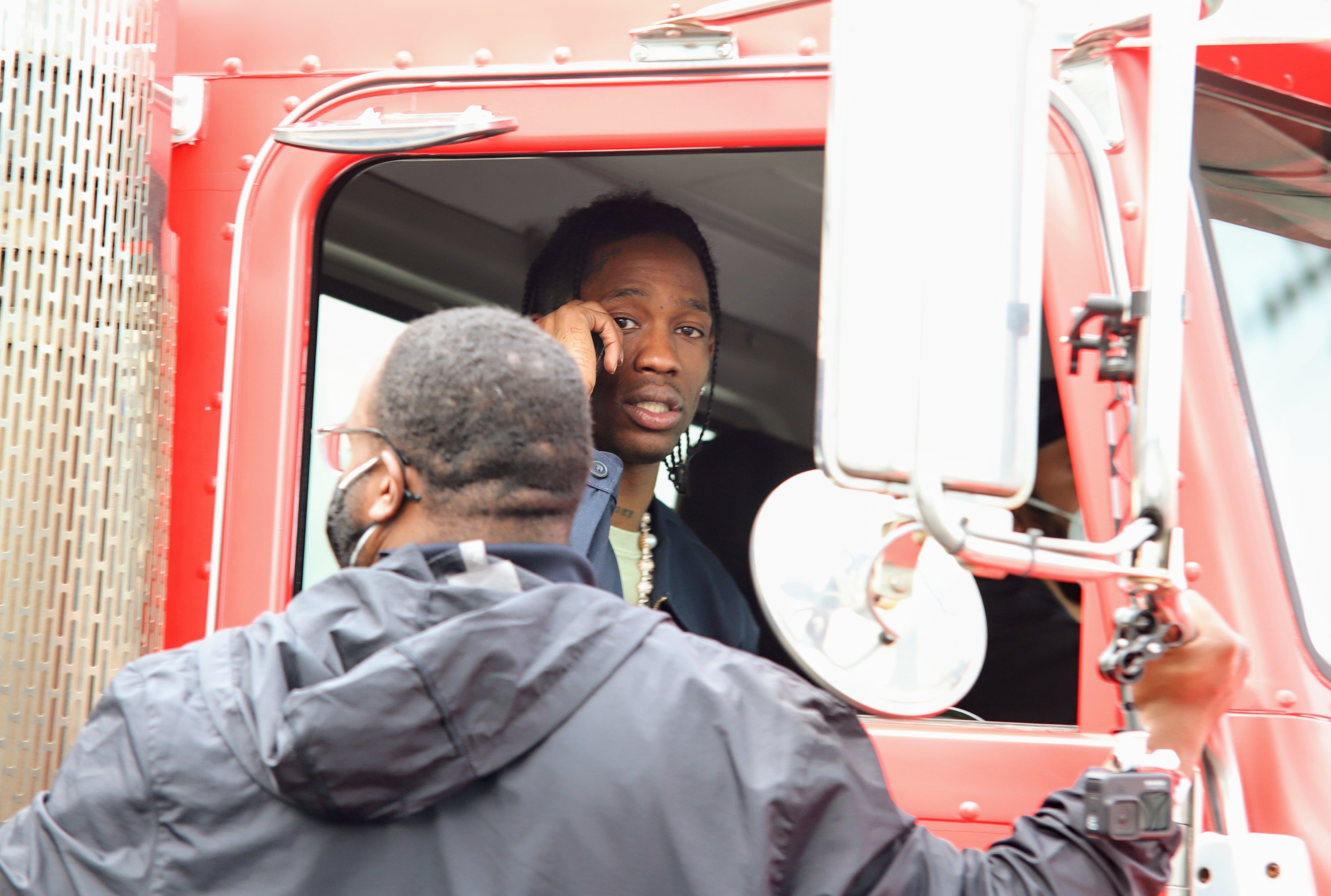 Travis Scott sits in the driver's seat of a truck while another man wearing glasses and a jacket speaks to him through the window