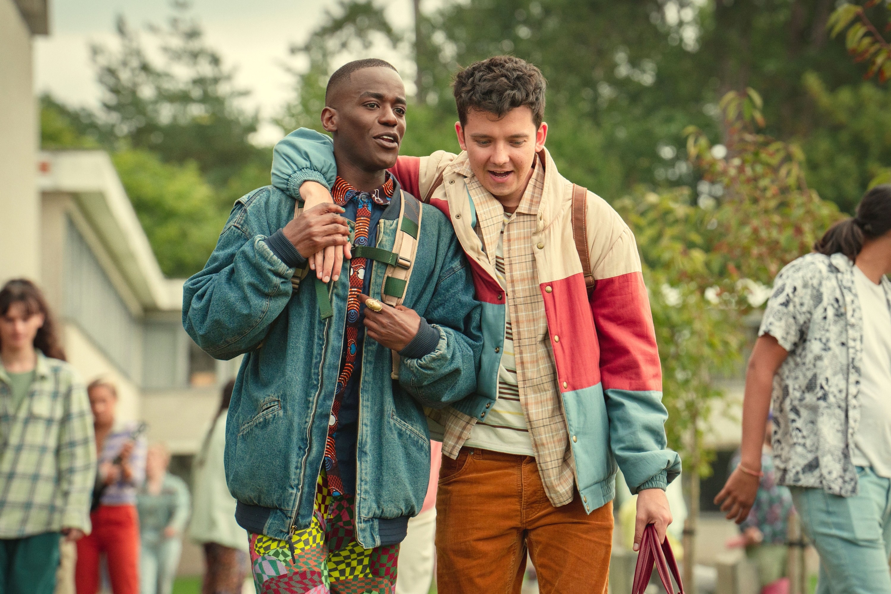 Ncuti Gatwa and Asa Butterfield, from "Sex Education," walk together in stylish jackets, laughing. Background shows other students on campus