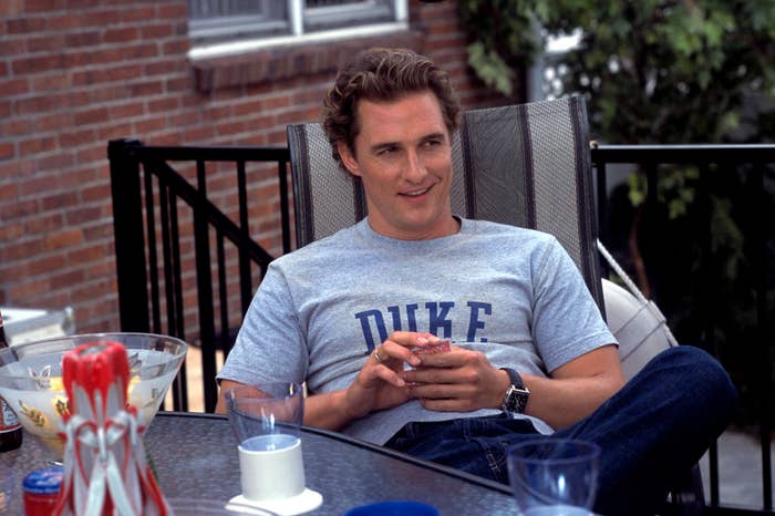 Matthew McConaughey sits in a chair on a patio, wearing a "Duke" t-shirt and jeans, holding a drink and smiling