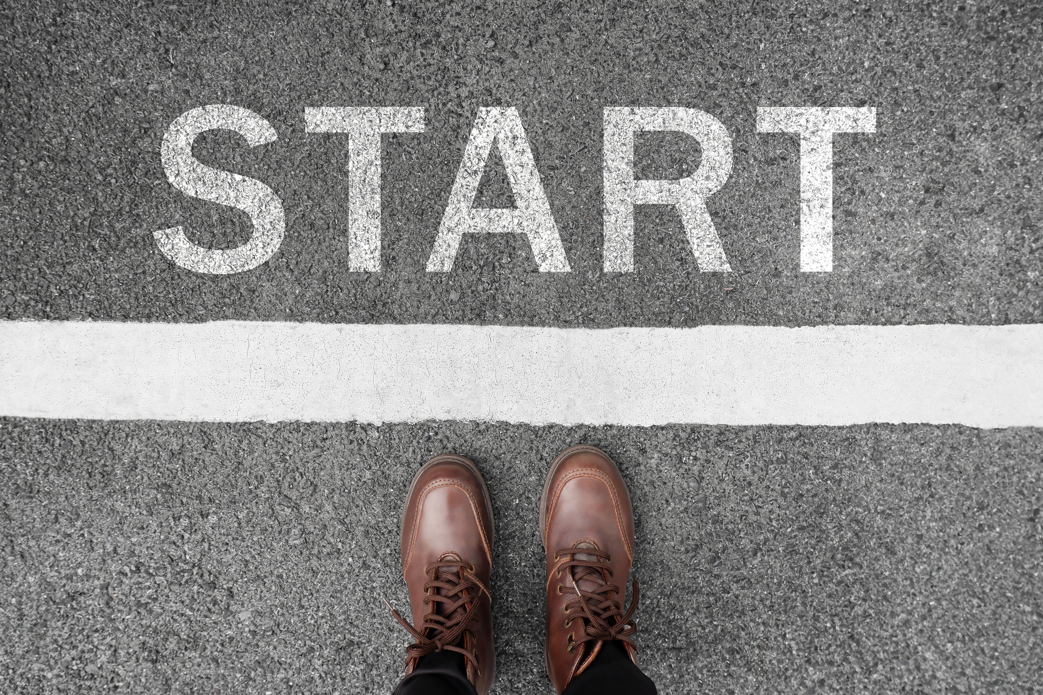 Person wearing brown shoes standing at a starting line on a paved surface with the word "START" written in large white letters on the ground