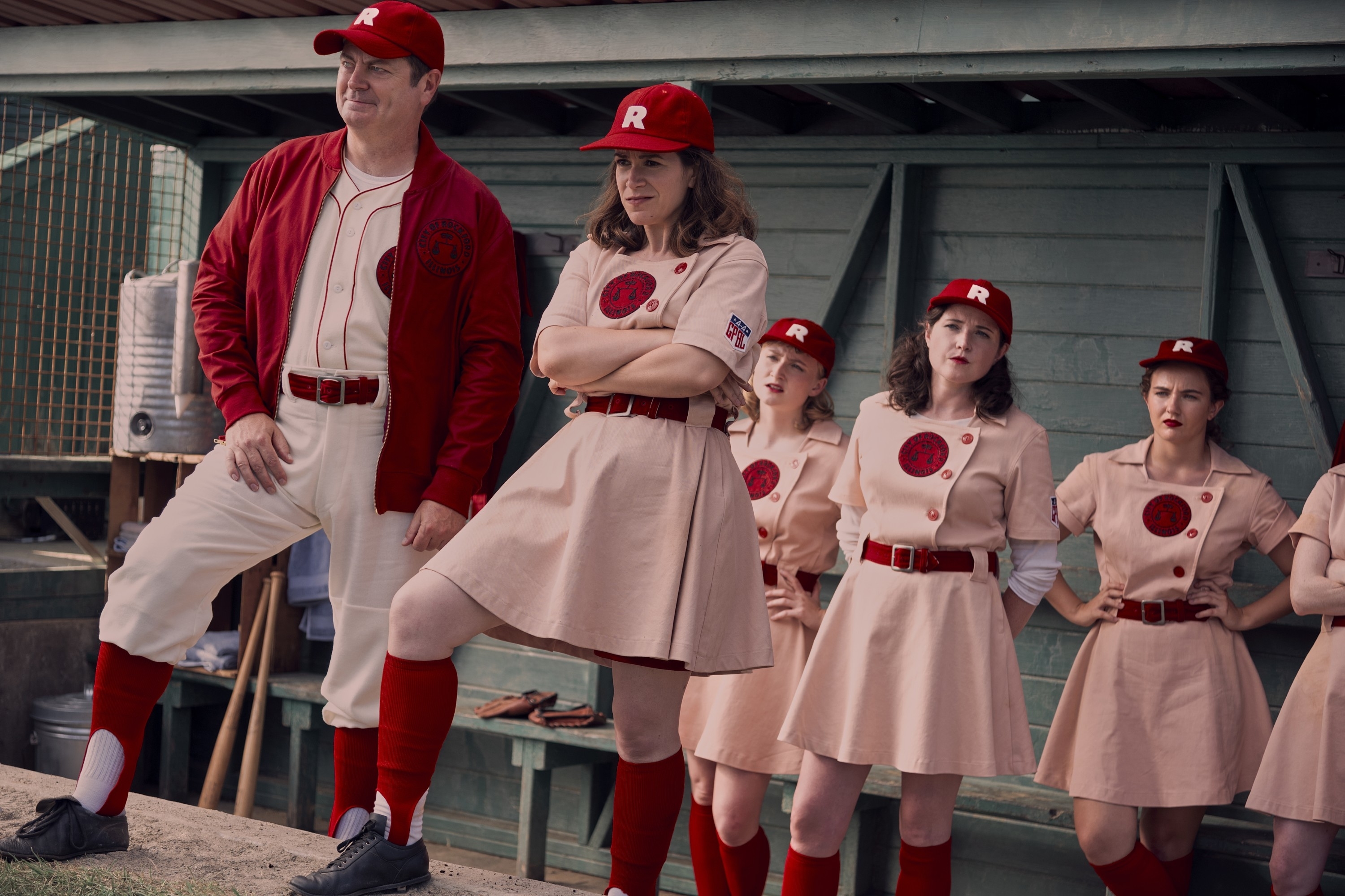 A League of Their Own TV series cast in baseball uniforms: Abby Jacobson, D'Arcy Carden, and other actresses stand confidently in a dugout