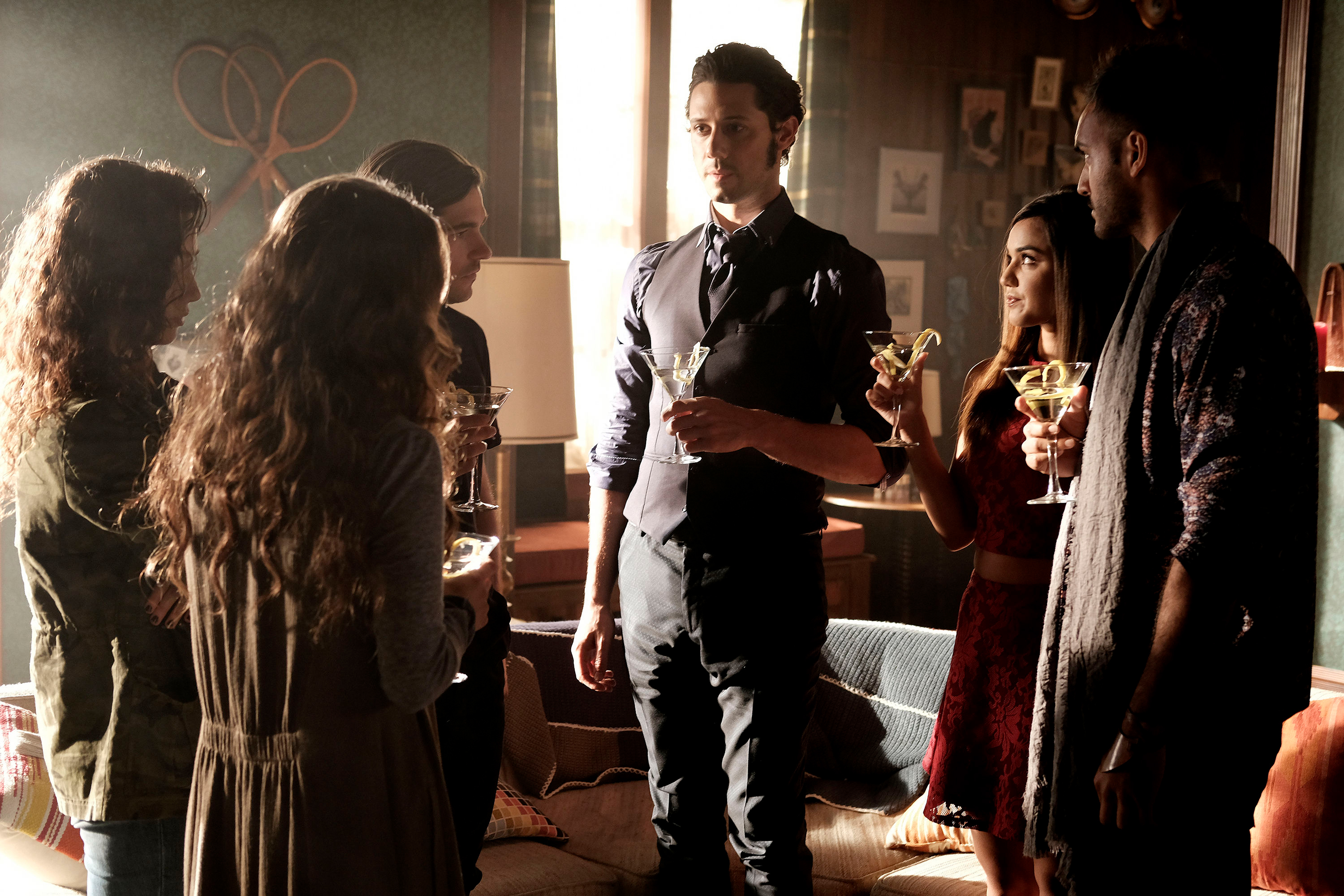 Group of actors from 'The Magicians' in a living room holding drinks, including Olivia Taylor Dudley, Stella Maeve, Hale Appleman, Summer Bishil, and Arjun Gupta