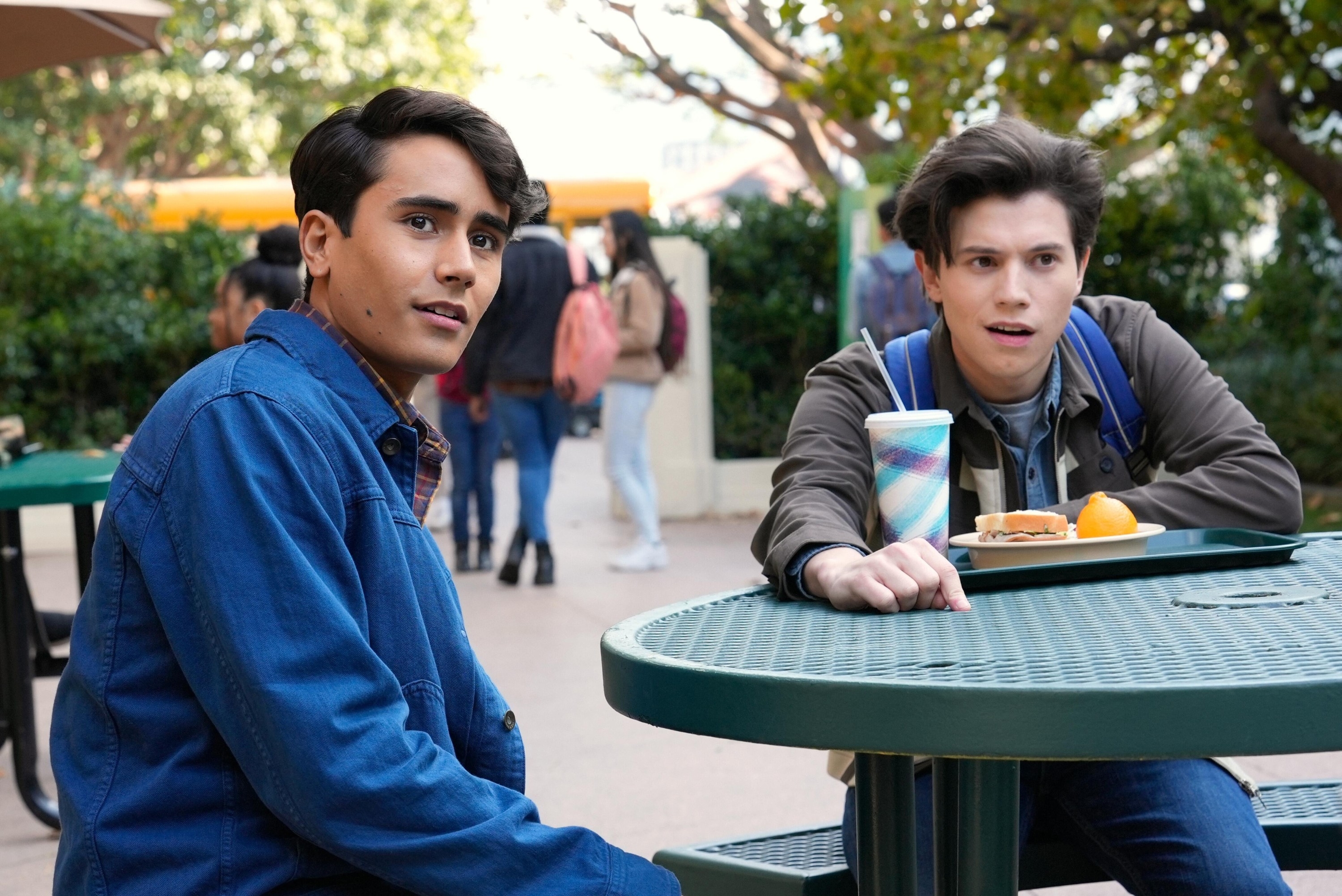 Michael Cimino and George Sear in a school courtyard scene from TV show "Love, Victor," seated at a picnic table with lunch trays