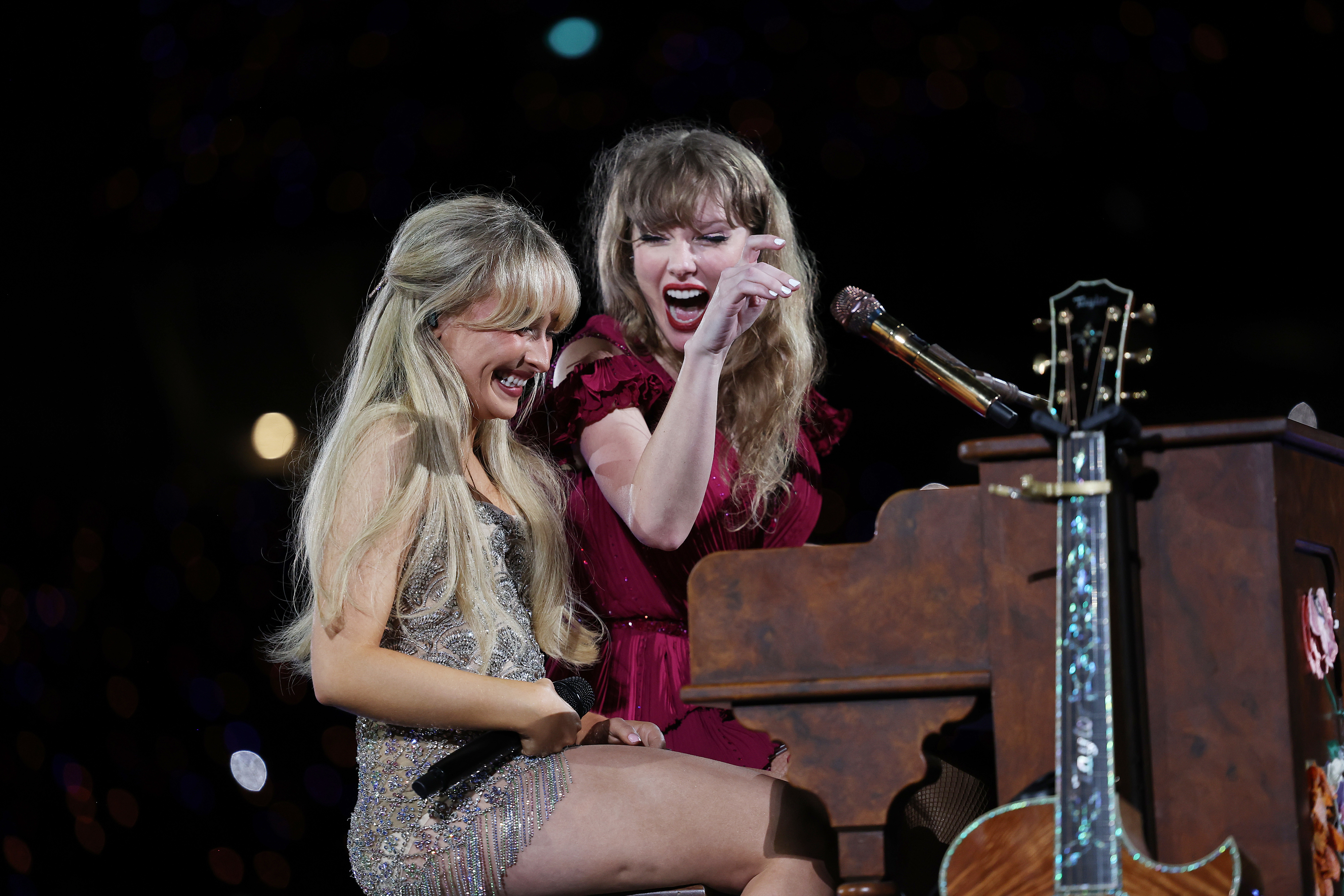 Sabrina Carpenter is in a sparkly dress, and Taylor Swift is in a red dress, joyfully performing at a piano on stage. Taylor Swift is pointing and smiling