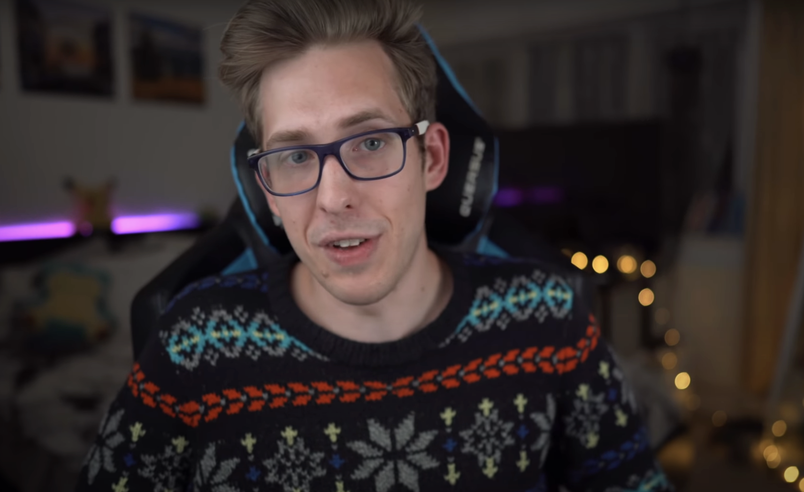 Evan Edinger is sitting in a room with a gaming chair, wearing glasses and a patterned sweater. The room has a cozy setup with string lights in the background