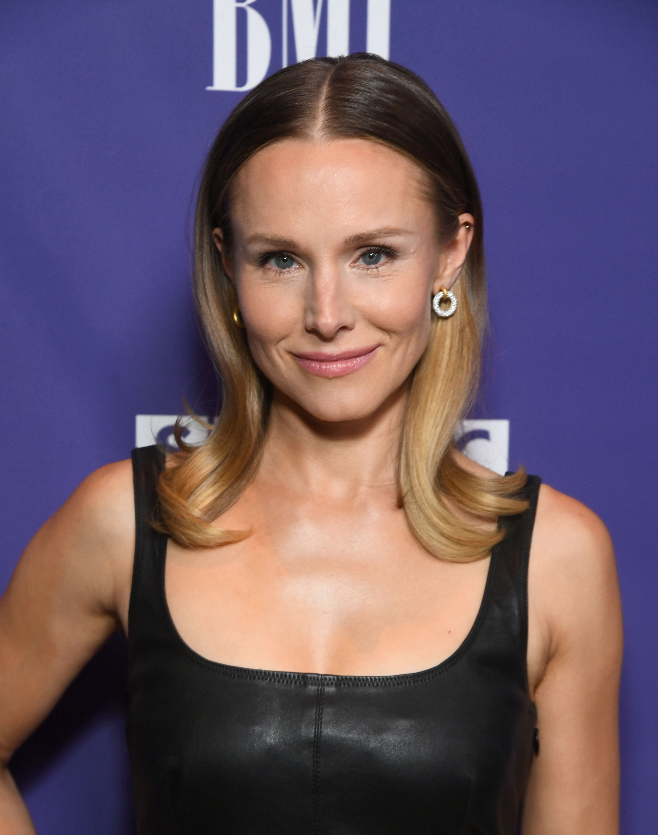 Kristen Bell at a BMI event, smiling and wearing a sleeveless leather dress with simple earrings