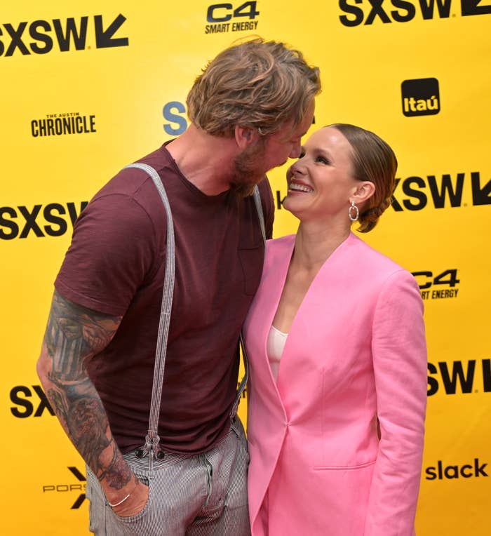 Kristen Bell in a pantsuit and Dax Shepard in a casual shirt and pants with suspenders, smiling at each other on the SXSW red carpet