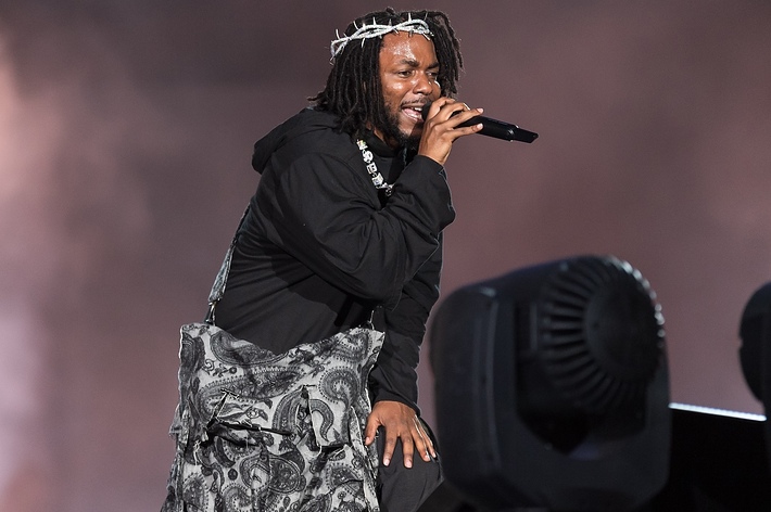 Kendrick Lamar performs on stage wearing a black outfit, a printed scarf, and a crown of thorns. He is singing into a microphone with a passionate expression