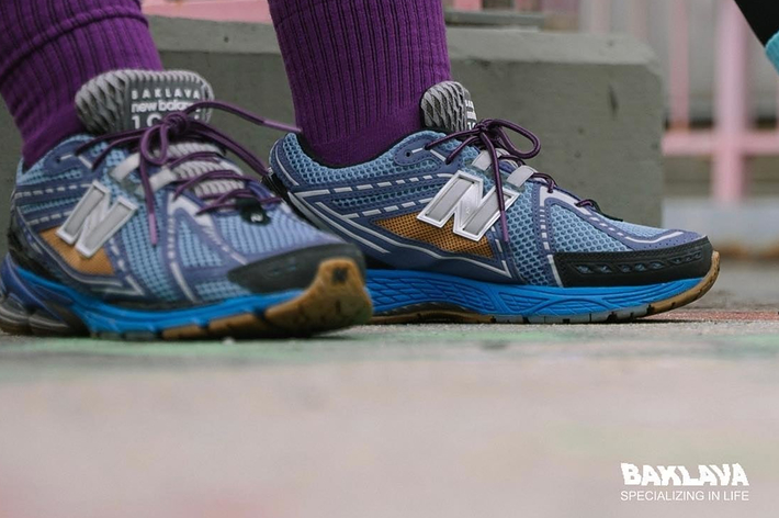 Two pairs of legs showing off New Balance sneakers: one person has purple socks and tattoos, the other wears black leggings and pink socks. Logos for Baklava and New Balance are visible
